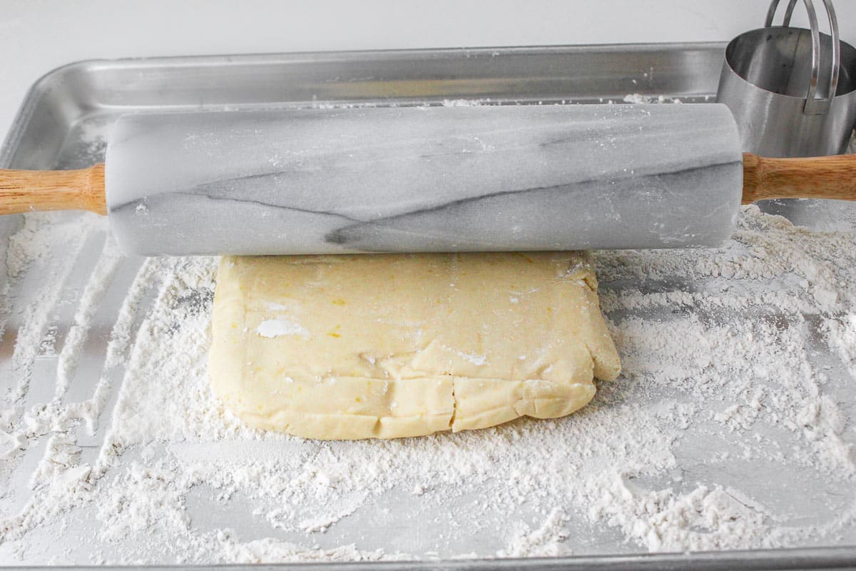 Cookie dough over flour being rolled out by rolling pin on baking sheet.