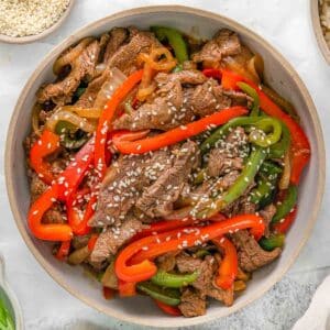 Thinly sliced strips of cooked steak, onion, red and green bell peppers topped with sesame seeds in a white bowl.