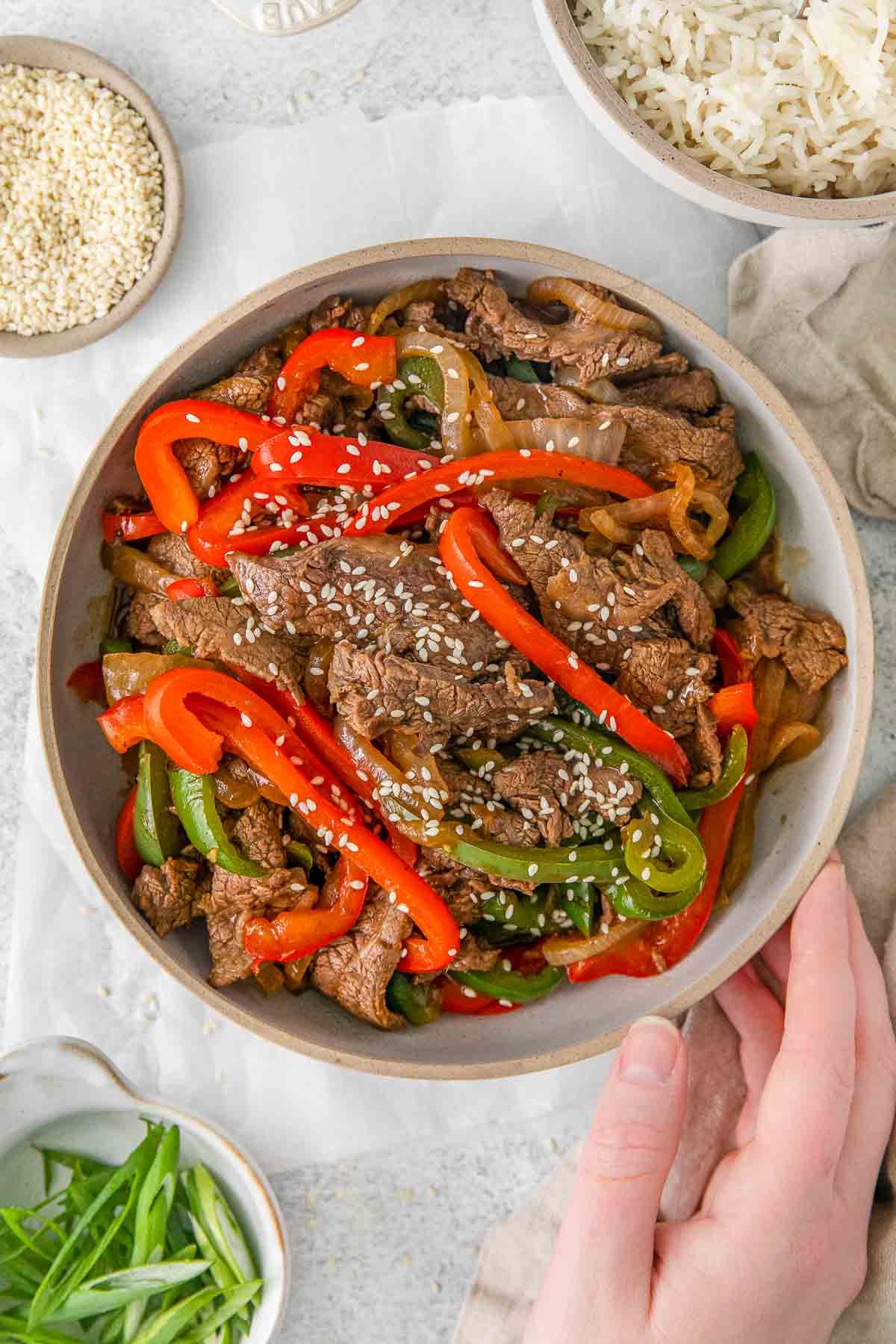 Cooked steak with onion, green and red bell peppers topped with sesame seeds in white bowl.