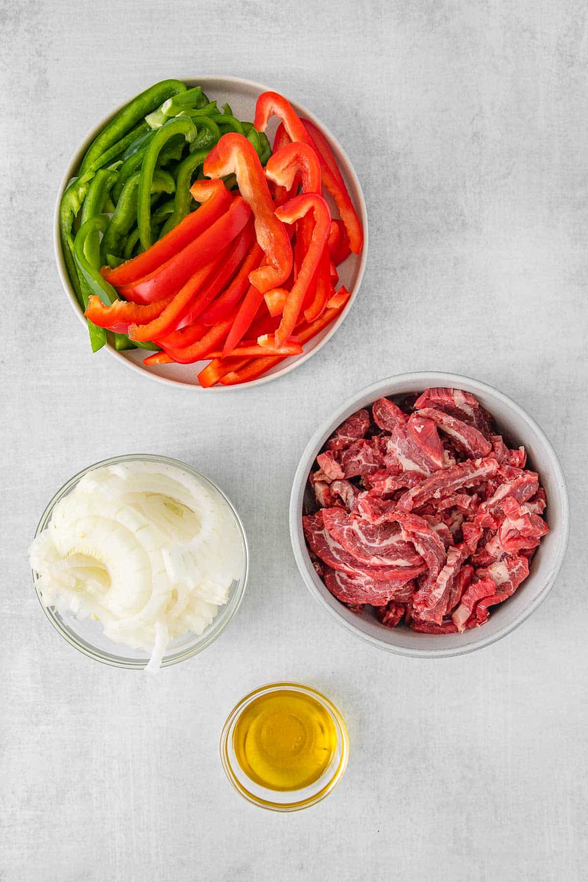 Several bowls with ingredients for Pepper Steak sauce - steak, olive oil, onion, green bell pepper and red bell pepper.