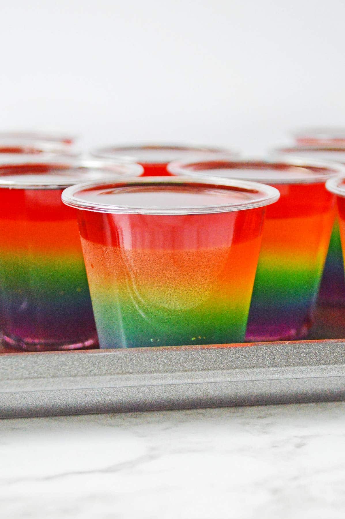 Plastic cups on sheet pan with strawberry jello layer added to rainbow jello shot.