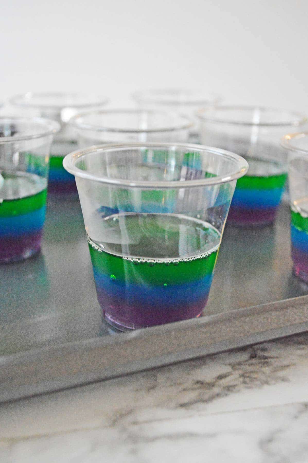 Plastic cups on sheet pan with lime jello layer added to rainbow jello shot.