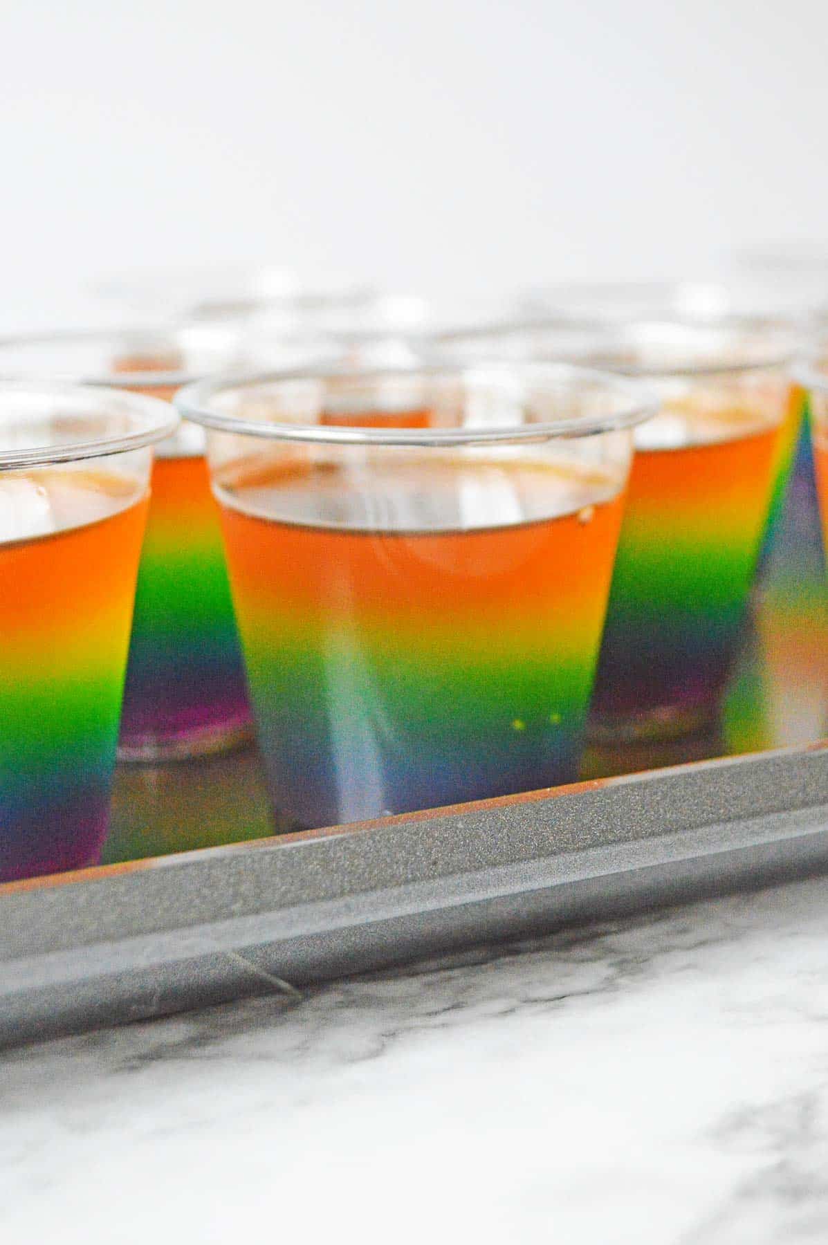 Plastic cups on sheet pan with orange jello layer added to rainbow jello shot.