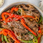 Thinly sliced strips of cooked steak, onion, red and green bell peppers topped with sesame seeds in a white bowl.