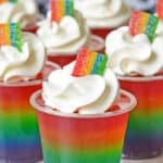 several rainbow jello shots with whipped cream and a rainbow colored candy on top.