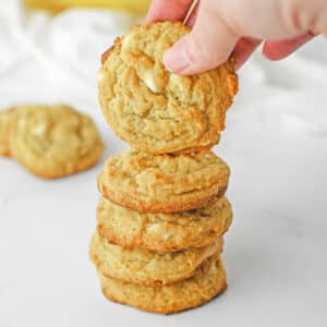 A stack of banana pudding cookies with white chocolate chips with one being held over the stack.