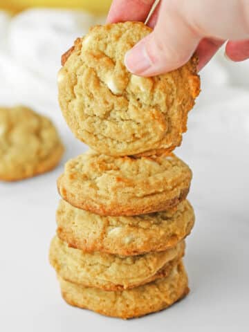 A stack of banana pudding cookies with white chocolate chips with one being held over the stack.