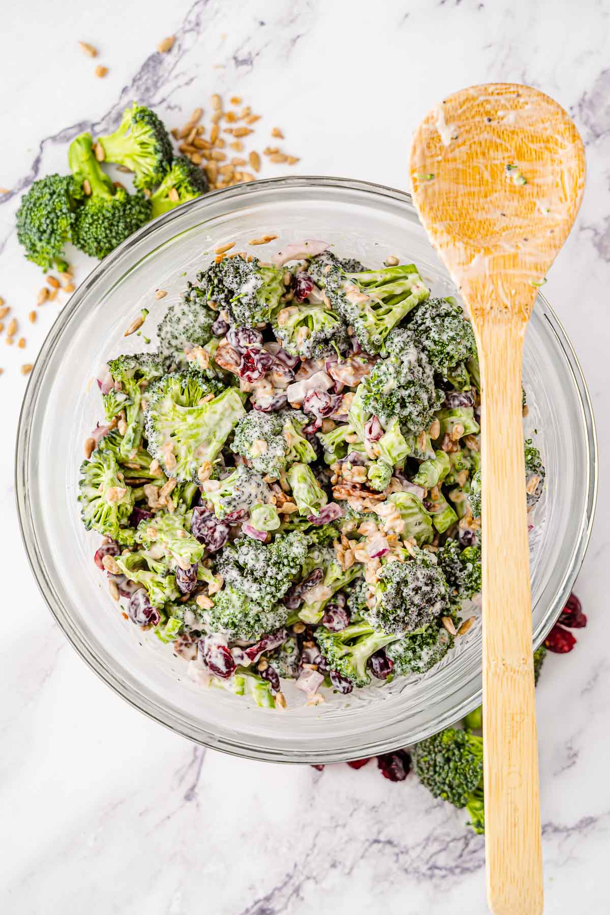A large glass bowl of broccoli salad ingredients being mixed together by a wooden spoon.