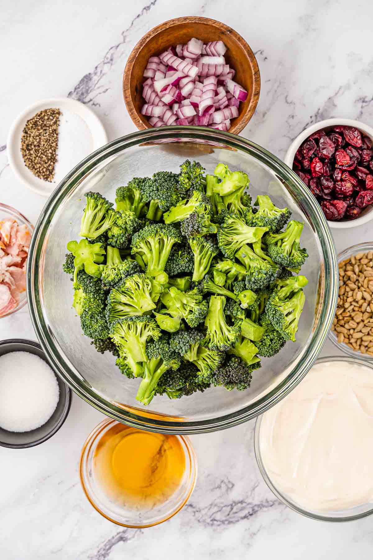 Several bowls of ingredients for broccoli salad - Apple cider vinegar, granulated sugar, mayonnaise, salt, black pepper, bacon bits, broccoli florets, dried cranberries, red onion diced, sunflower seeds.