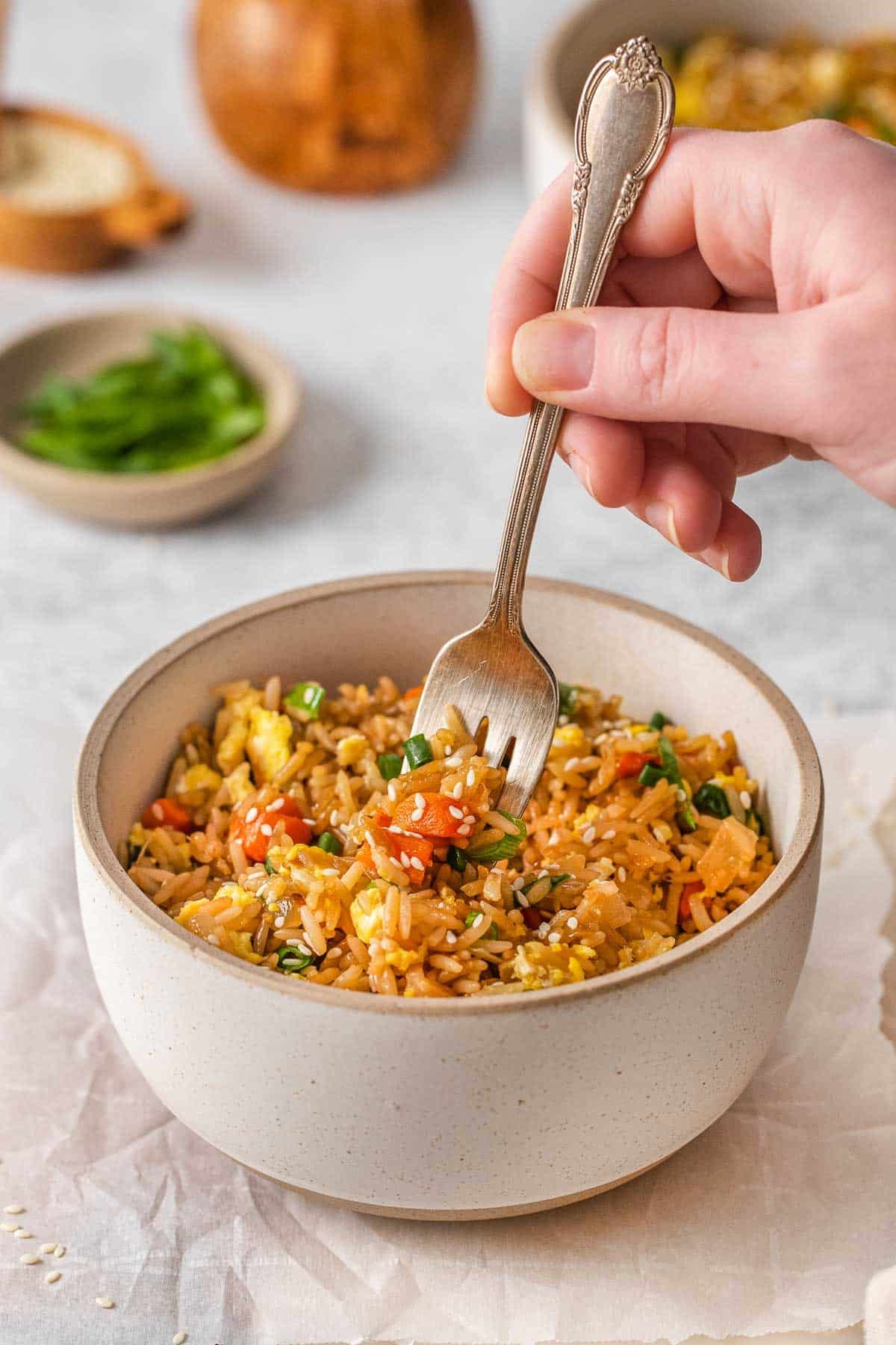 Fork held by womans hand being inserted into egg fried rice in a white bowl.