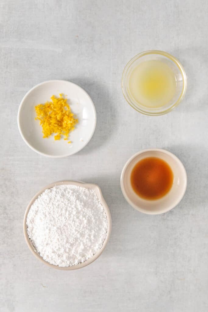 Several white bowls of ingredients for lemon glaze - Powdered sugar, lemon juice, lemon zest and vanilla extract.