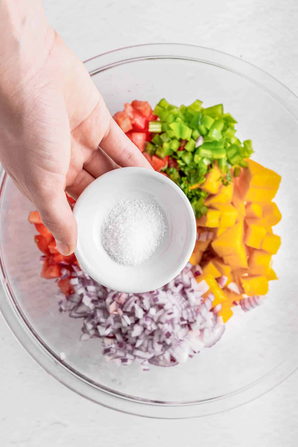 Small bowl of salt being held over large bowl of mango salsa ingredients.