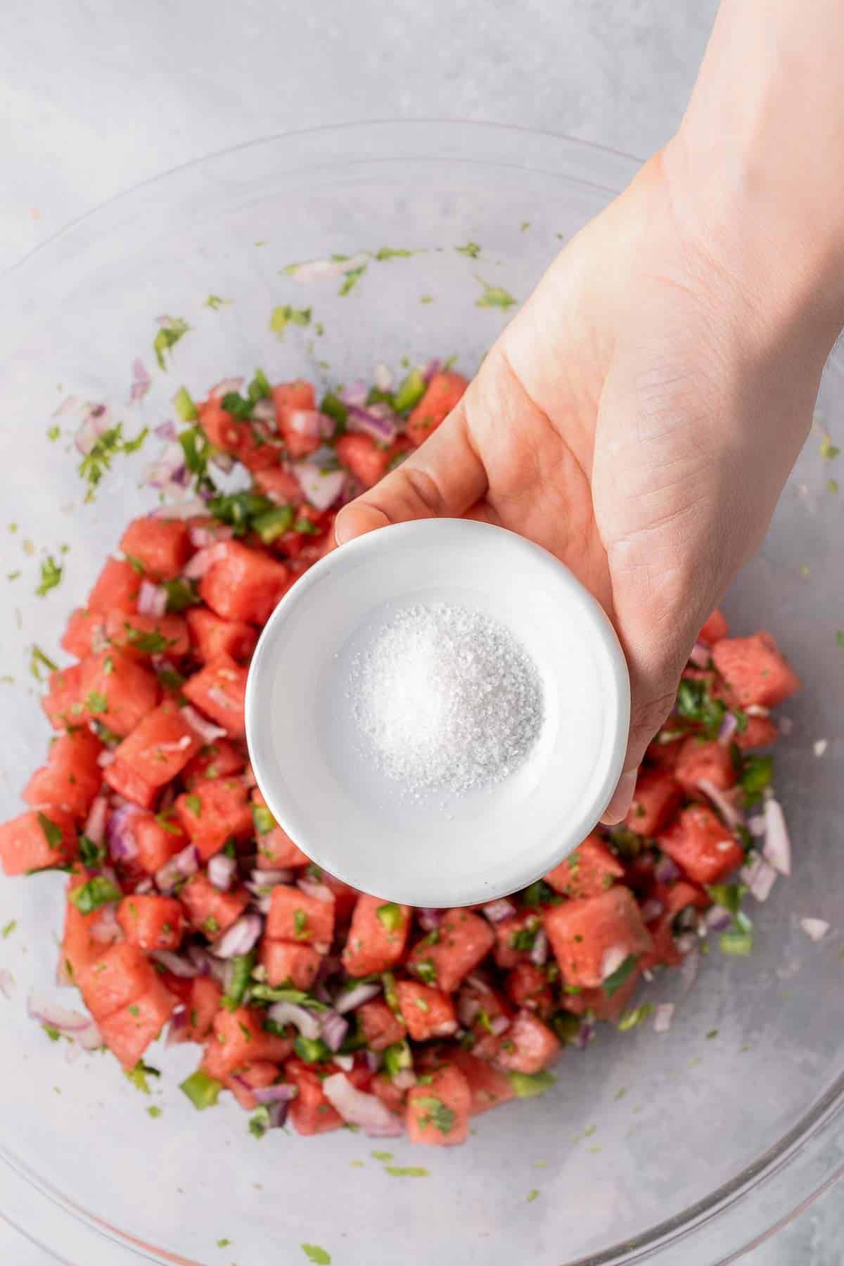 Small bowl of salt held over large glass bowl of watermelon salsa.