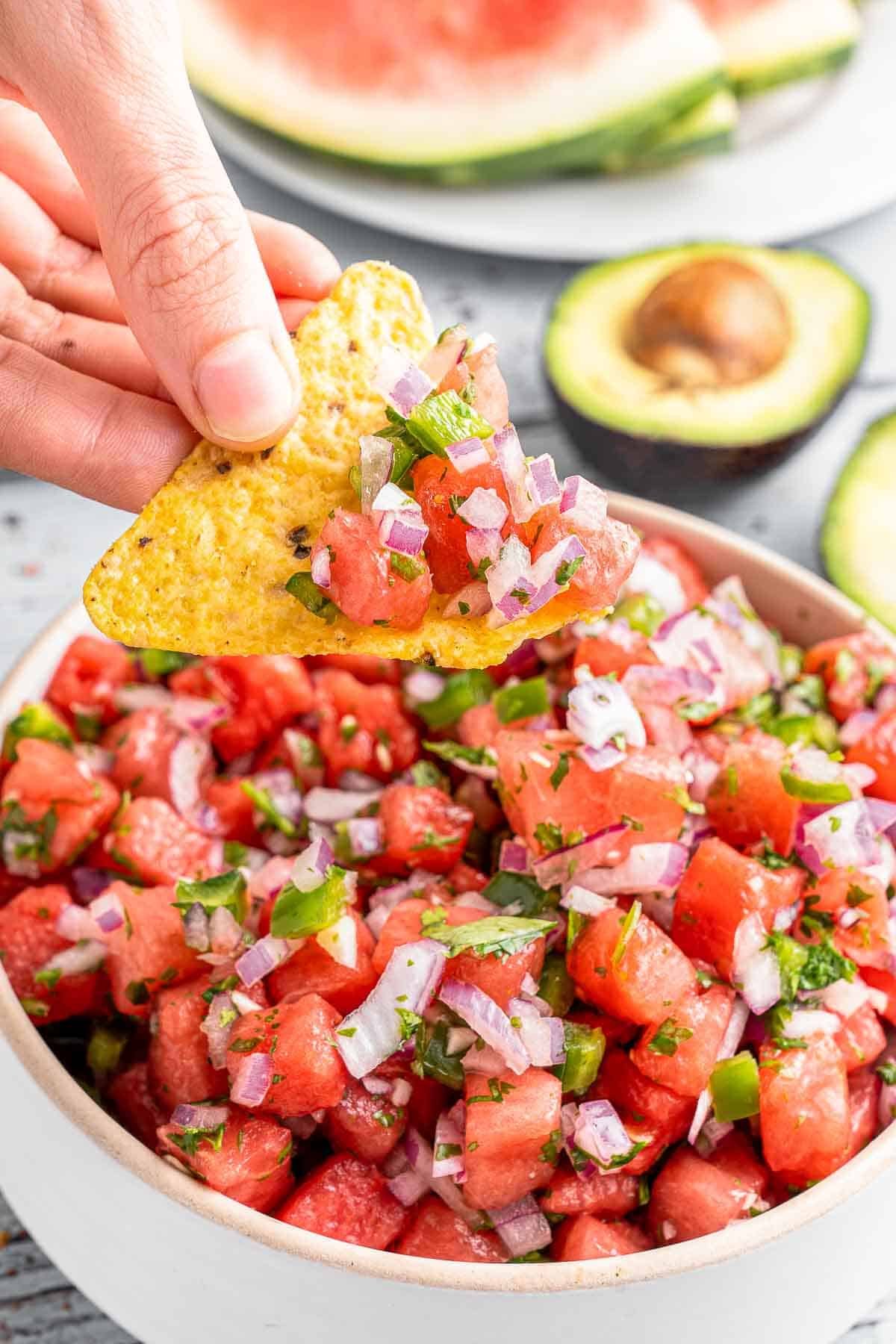 Tortilla chip scooping watermelon salsa out of white bowl.