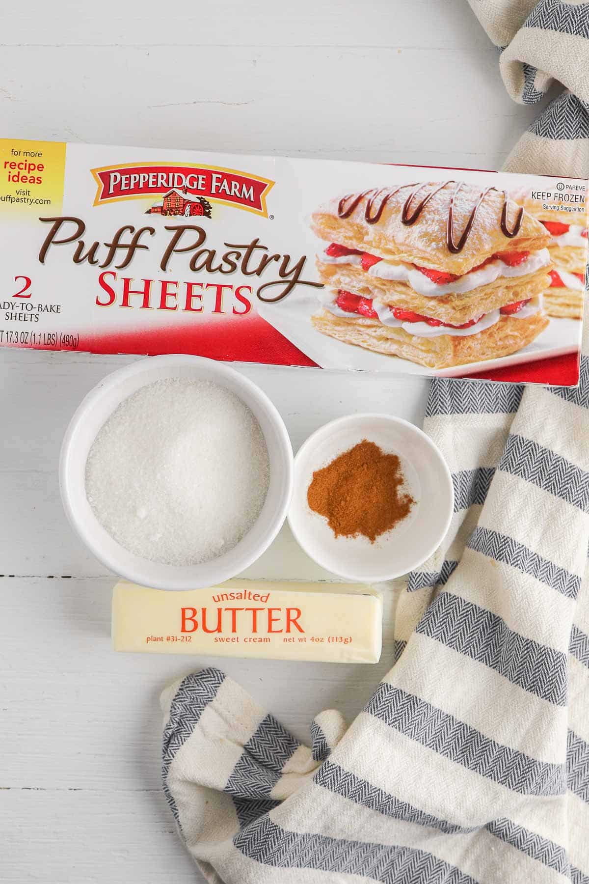 box of pepperidge farm puff pastry sheets, stick of butter, a small white bowl of sugar and a small bowl of ground cinnamon.