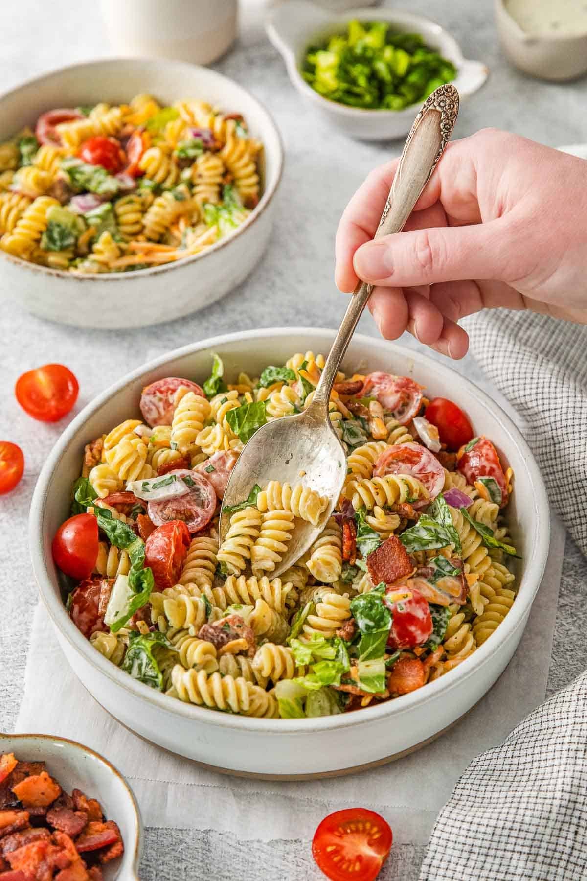 Hand holding spoonful of BLT pasta salad in a white bowl.