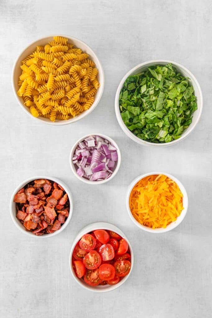 Several bowls of ingredients for BLT pasta salad - rotini pasta, romaine lettuce, cherry tomatoes, bacon, shredded cheddar cheese, red onion.
