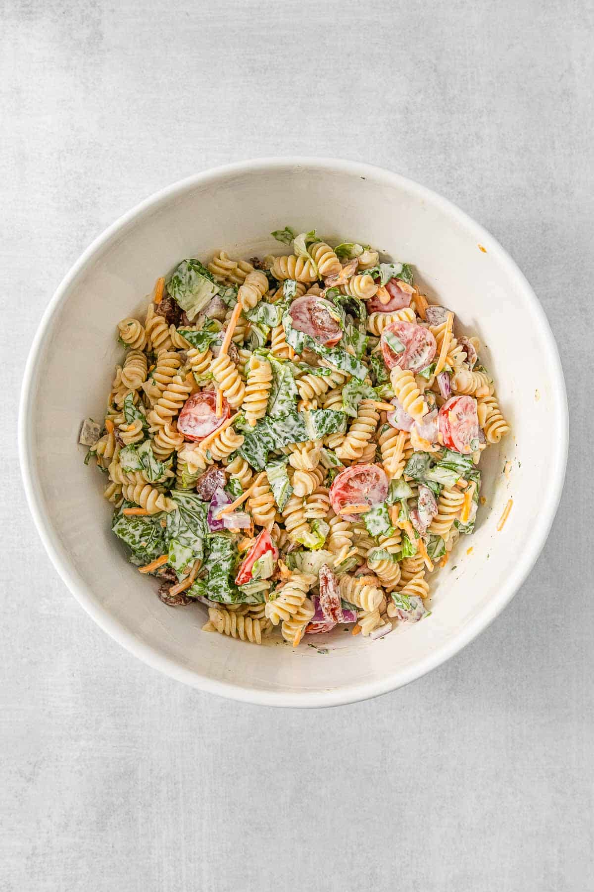 Pasta salad ingredients mixed with salad dressing ingredients in a large white mixing bowl.
