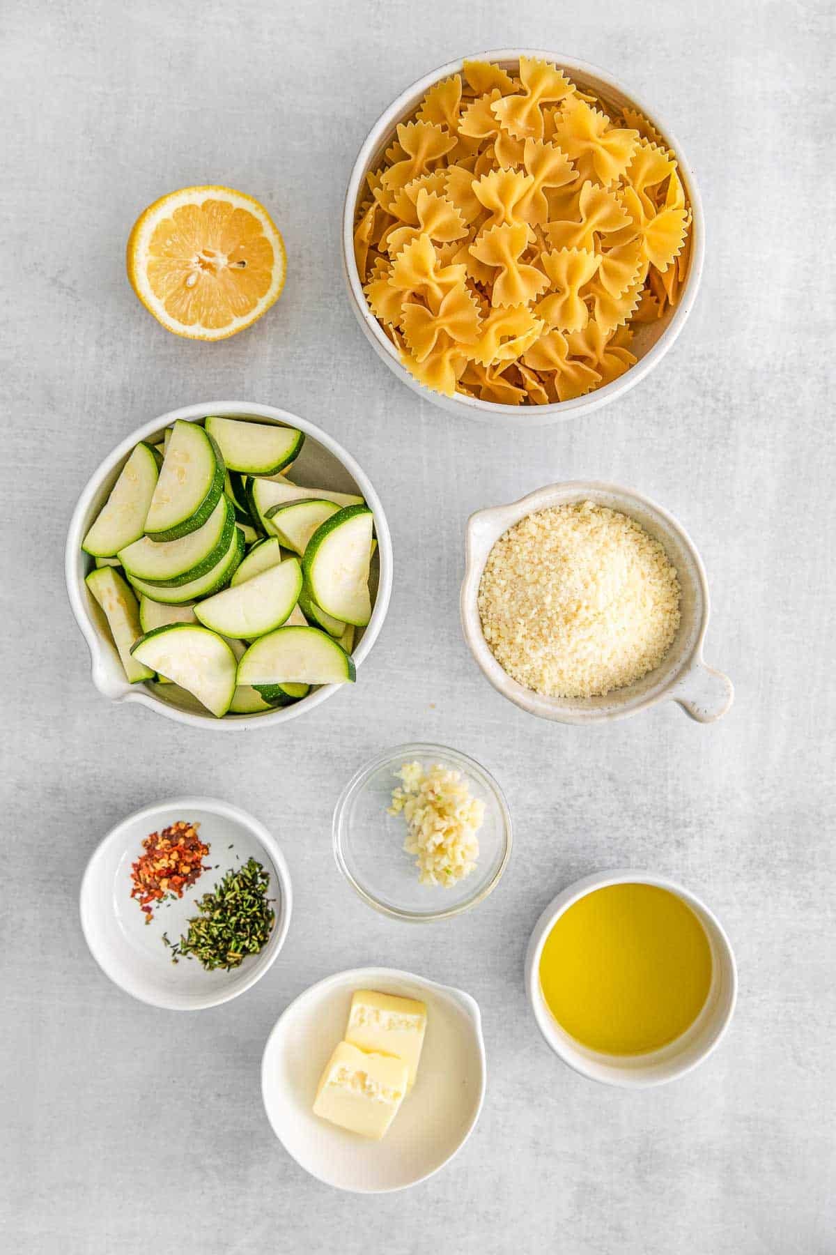 Several bowls of ingredients for lemon zucchini pasta - Bowtie pasta, olive oil, zucchini, garlic, thyme, crushed red pepper, butter, lemon, parmesan cheese, fresh parsley and toasted pine nuts.