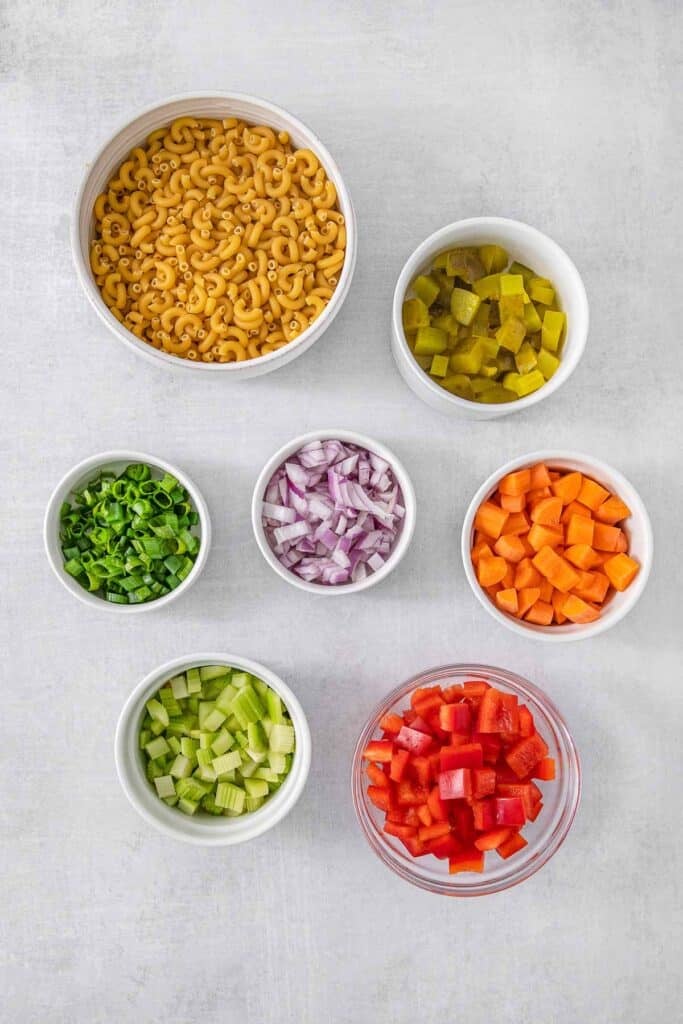Several white bowls of ingredients for Macaroni Salad - macaroni, red bell pepper, carrot, celery, dill pickles, red onion and green onions.