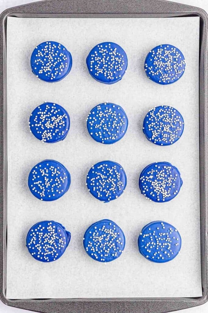 Multiple blue patriotic oreos with white sprinkles on a baking sheet.