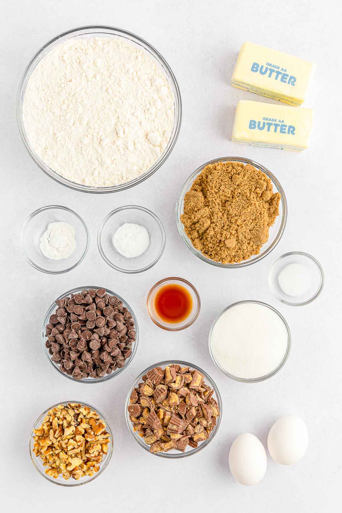 several glass bowls with ingredients for peanut butter cup cookie bars - flour, brown sugar, white sugar, vanilla, eggs, chocolate chips, peanut butter cups and chopped nuts.