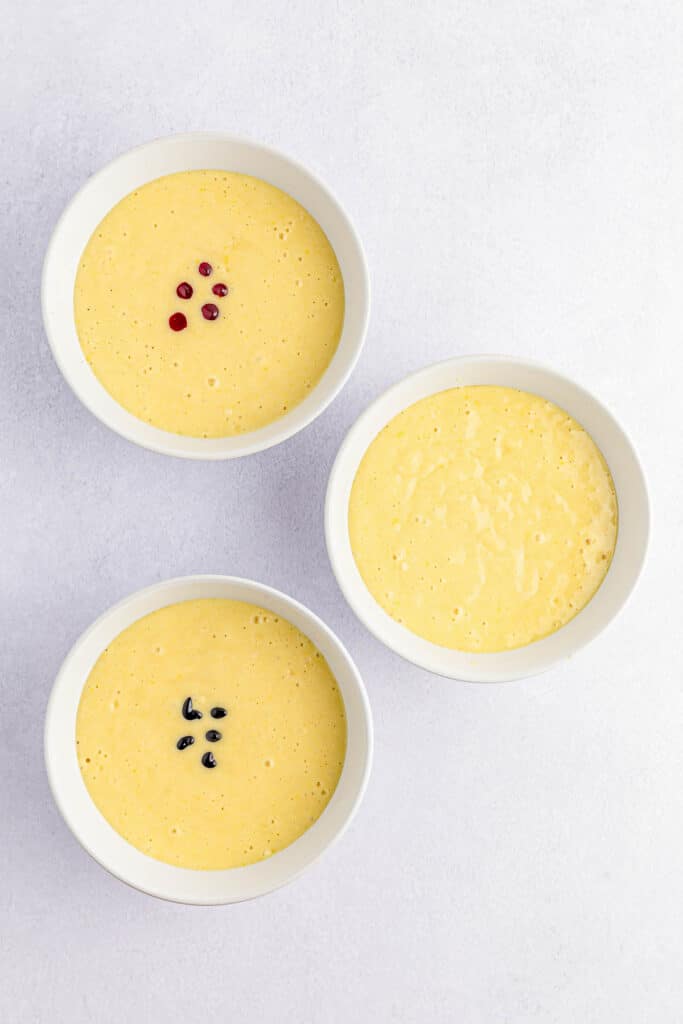 three small white bowls of cake mix. two with 5 drops of food color.