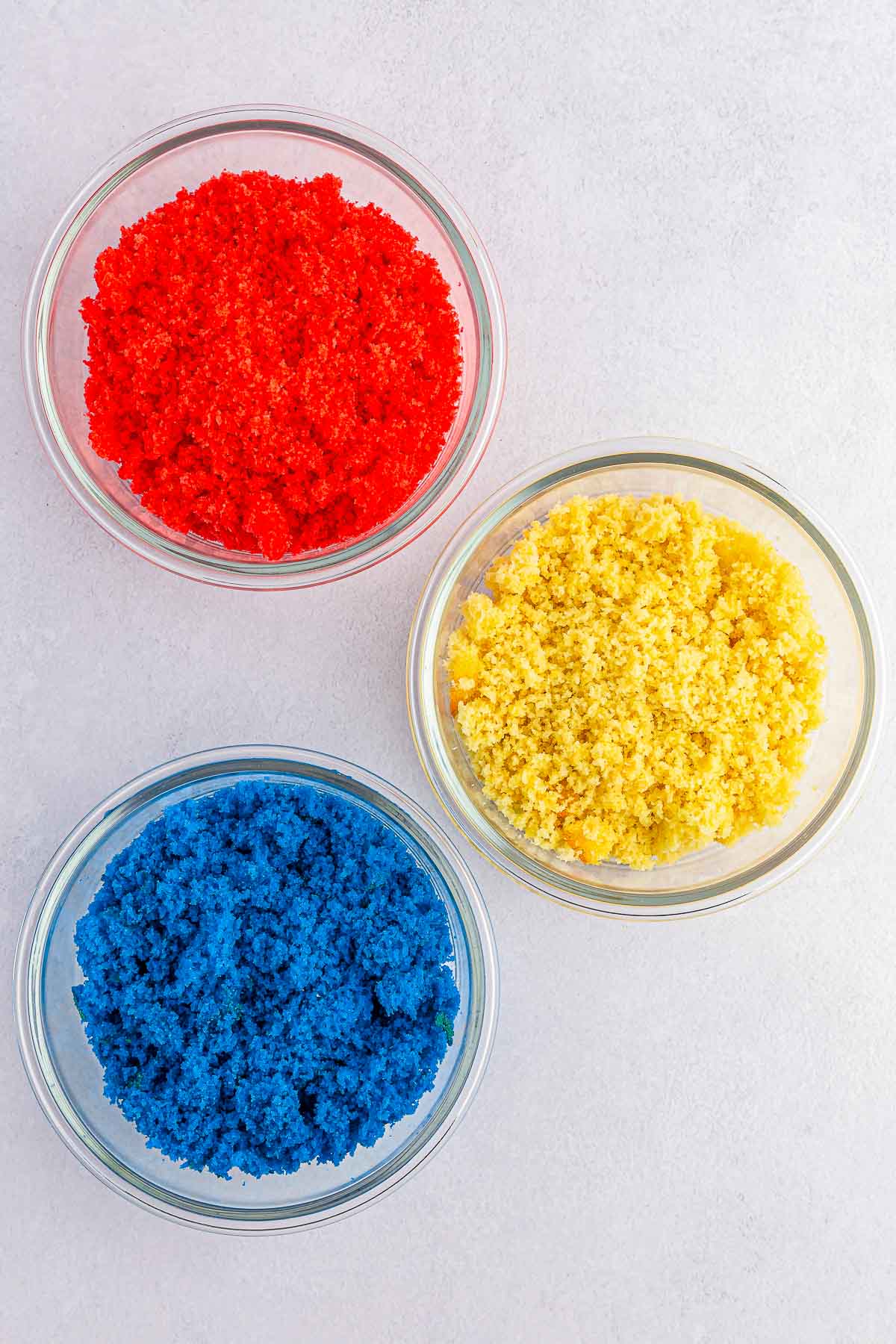 three glass bowls of crumbled cake batter. one blue, one red and one white.
