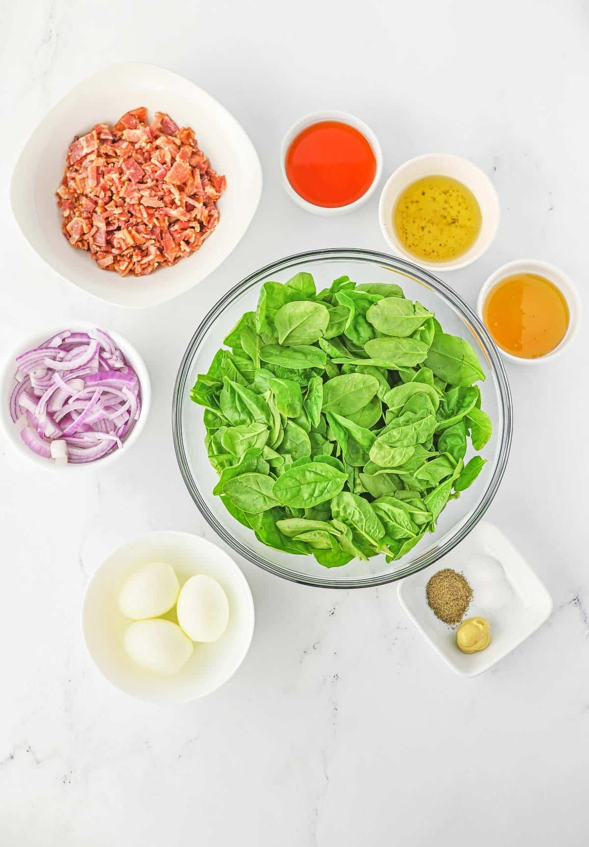 Several bowls of ingredients for spinach salad - Baby Spinach, bacon, bacon grease, red onion, eggs, red wine vinegar, honey, dijon mustard, salt and pepper.