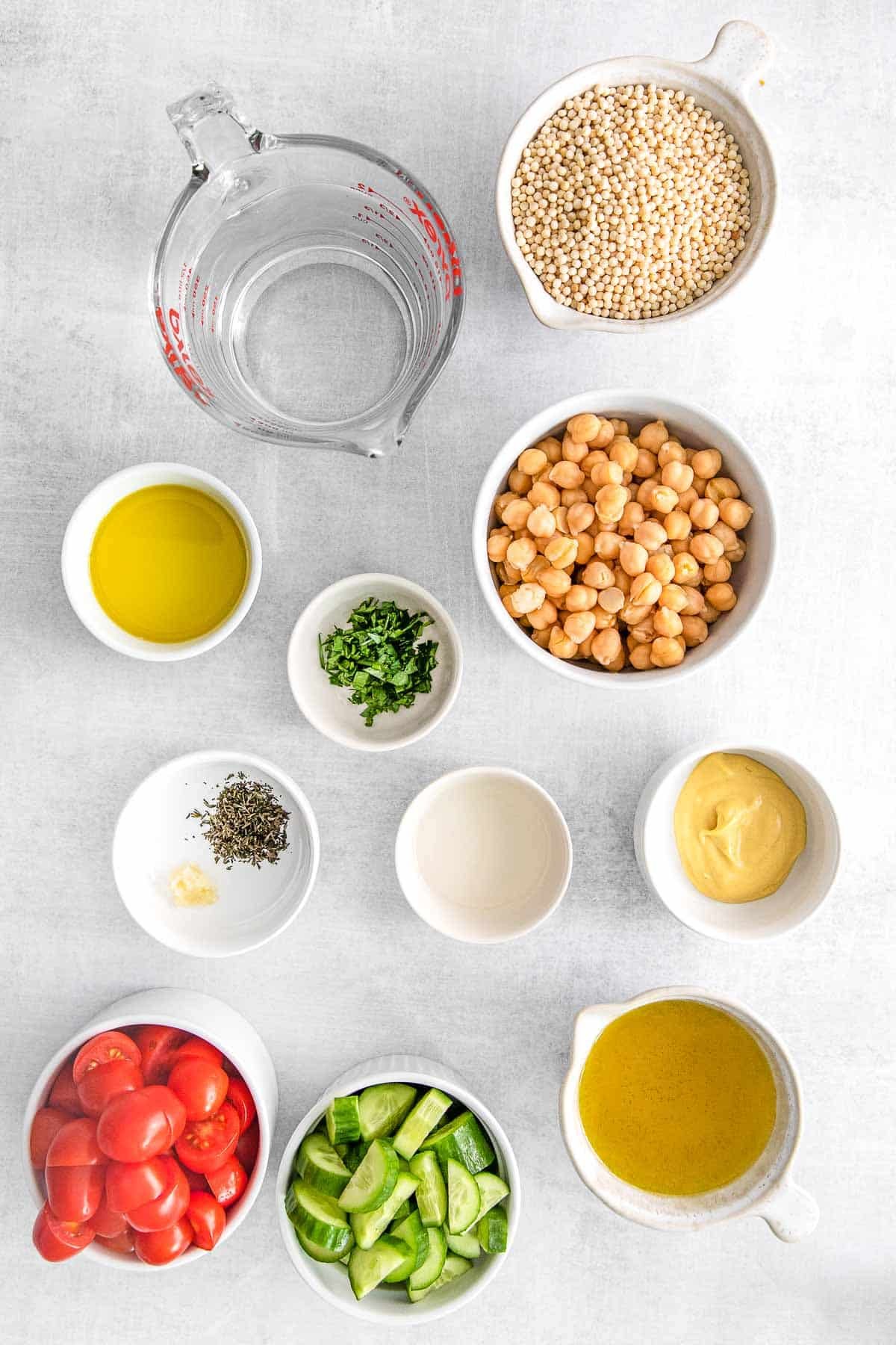 Several bowls of ingredients for Mediterranean couscous salad - Olive oil, Pearl couscous, Water, Chickpeas, Persian cucumbers, Cherry tomatoes, White wine vinegar, Dijon mustard, Fresh parsley, Garlic clove, Fresh thyme, Olive oil.