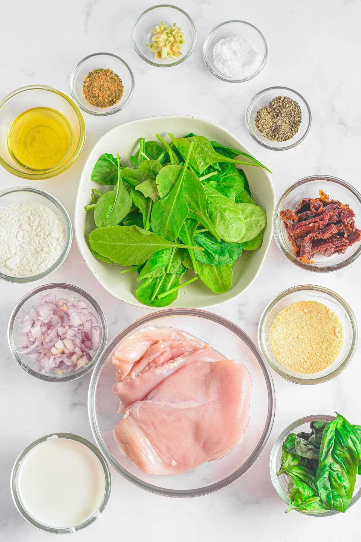 several glass bowls with ingredients for tuscan chicken. raw chicken breasts, baby spinach, sun dried tomatoes, red onion, oil, cream, parmesan cheese, garlic, and spices.