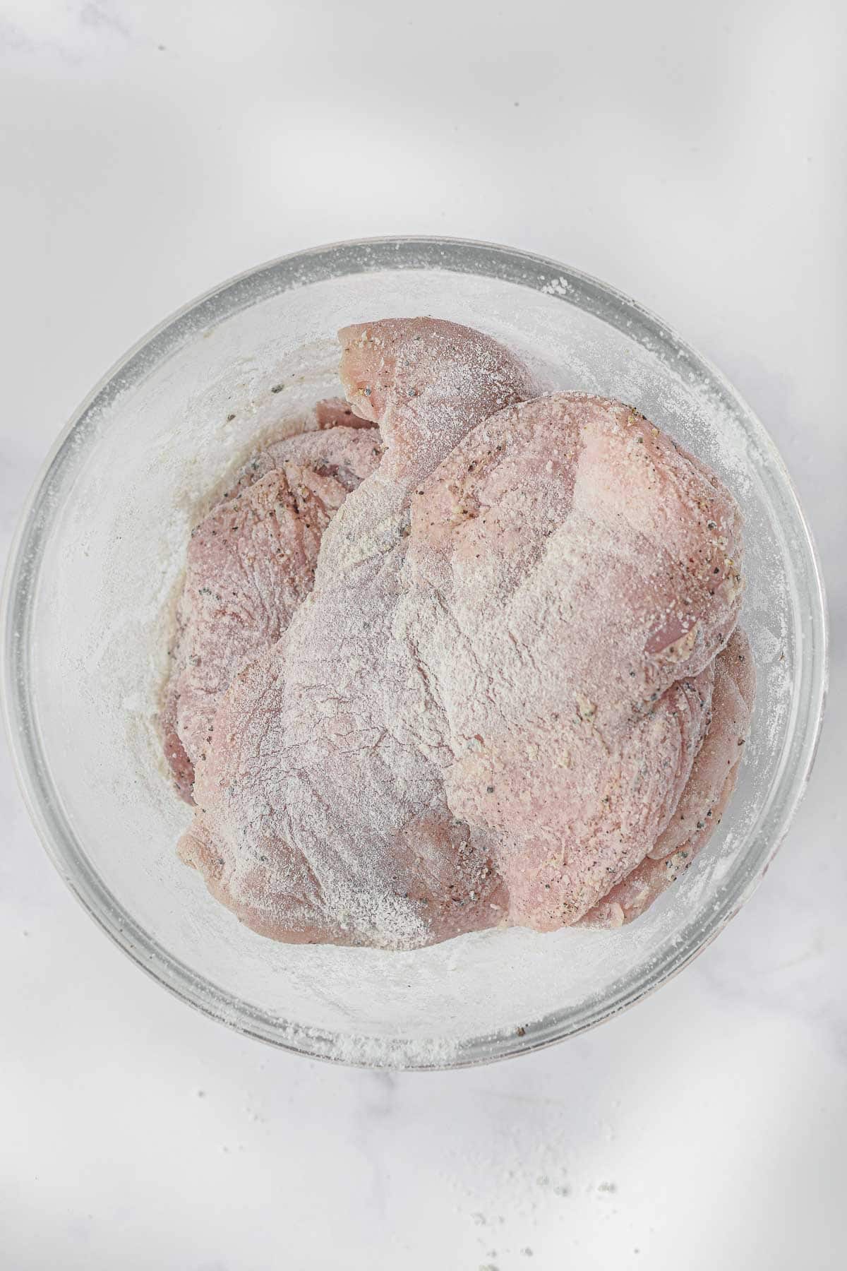 glass mixing bowl with raw chicken breasts dredged in white flour.