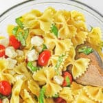 Caprese Pasta Salad with mozzarella balls, cherry tomatoes and fresh basil in a large glass bowl with wooden spoon in it.