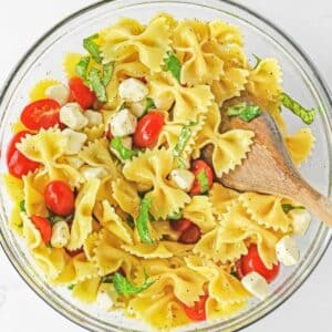 Caprese Pasta Salad with mozzarella balls, cherry tomatoes and fresh basil in a large glass bowl with wooden spoon in it.