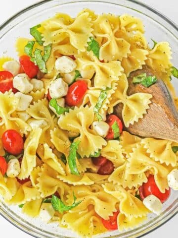 Caprese Pasta Salad with mozzarella balls, cherry tomatoes and fresh basil in a large glass bowl with wooden spoon in it.