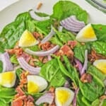 Spinach salad with bacon, hard boiled eggs and sliced red onions tossed in a bacon dressing in a white bowl.
