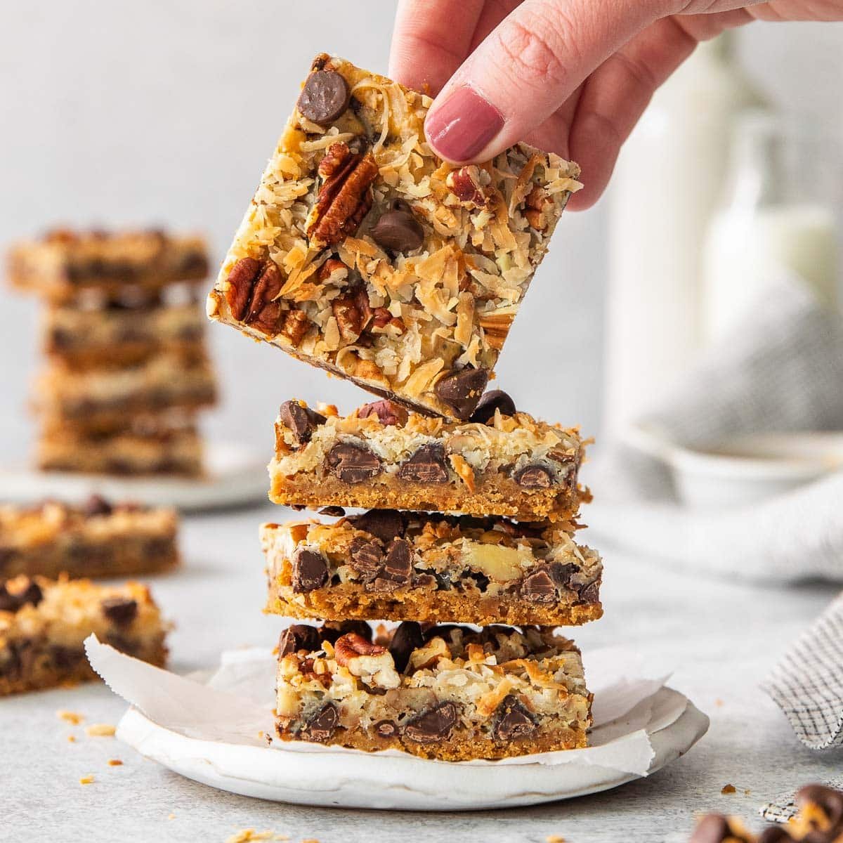 Stack of 4 magic cookie bars on a white plate with top one being picked up by woman's hand.