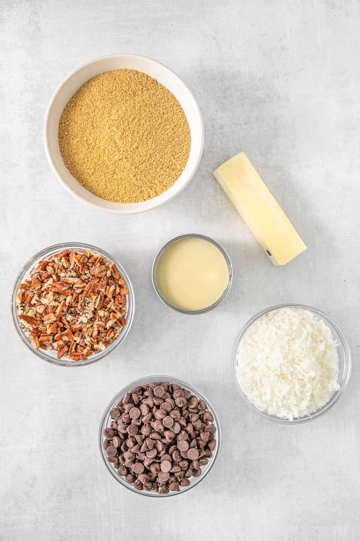 Several bowls of ingredients for Magic Cookie Bars - Graham Cracker Crumbs, Salted Butter, Sweetened Condensed Milk, Dark Chocolate Chips, Chopped Pecans and Shedded Sweetened Coconut.