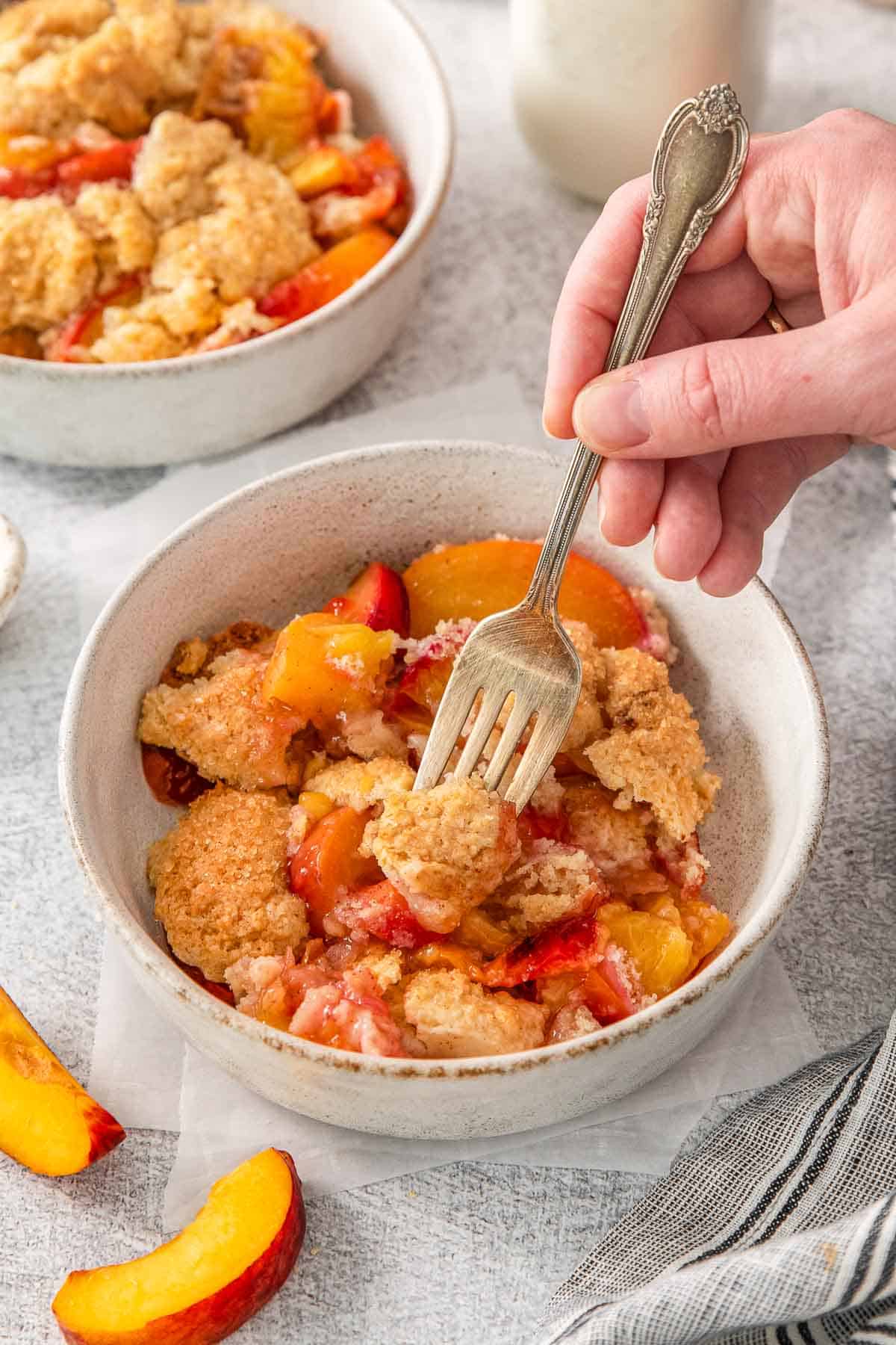 Peach cobbler in a white bowl with hand holding fork in dish.