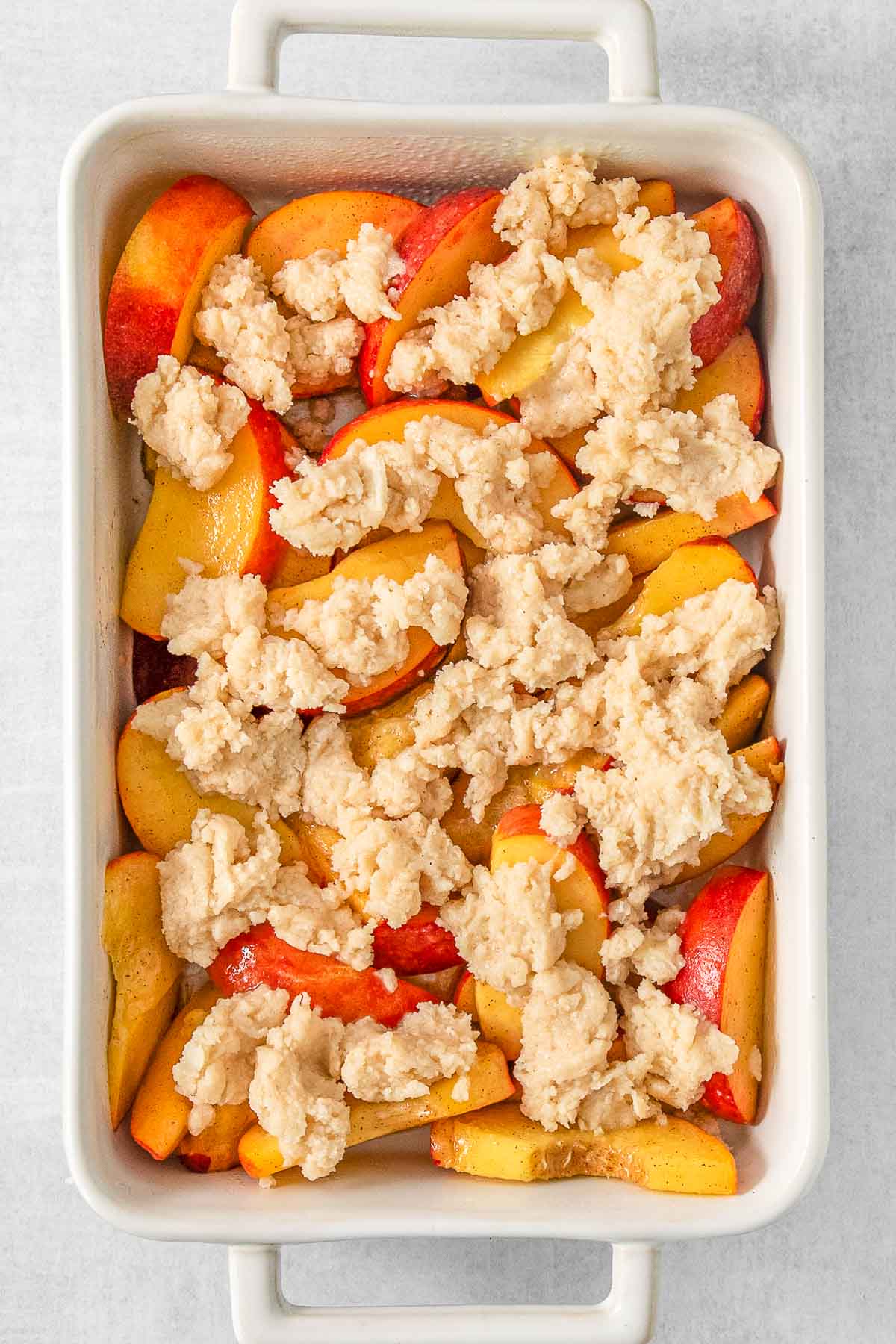 Baking dish full of sliced peaches and cobbler topping sprinkled overtop.