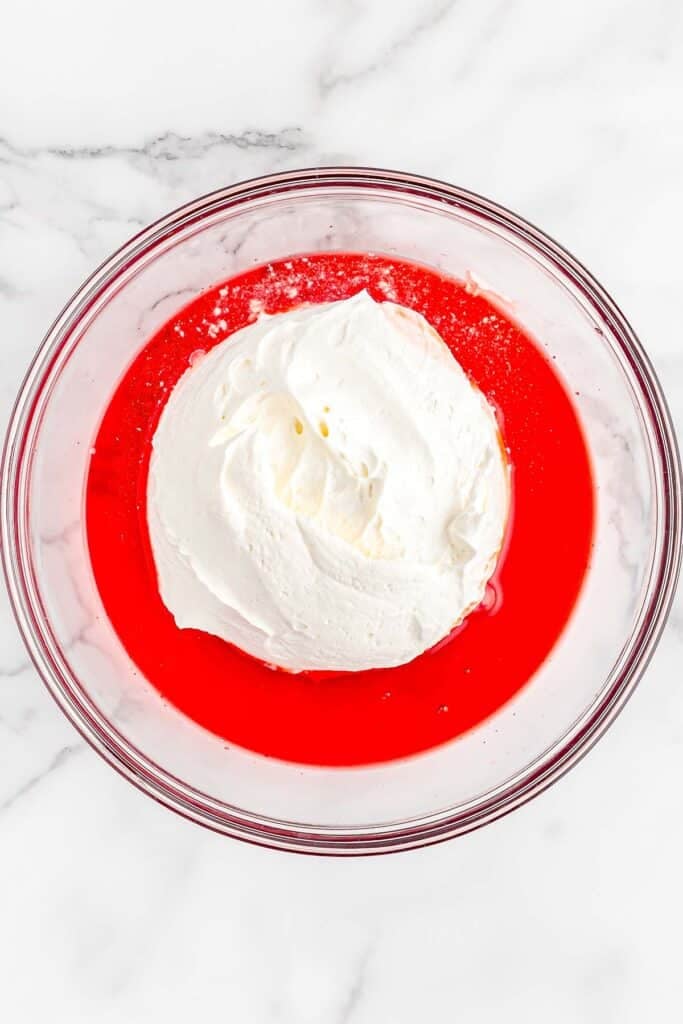Cool whip and strawberry jello mix in glass mixing bowl.
