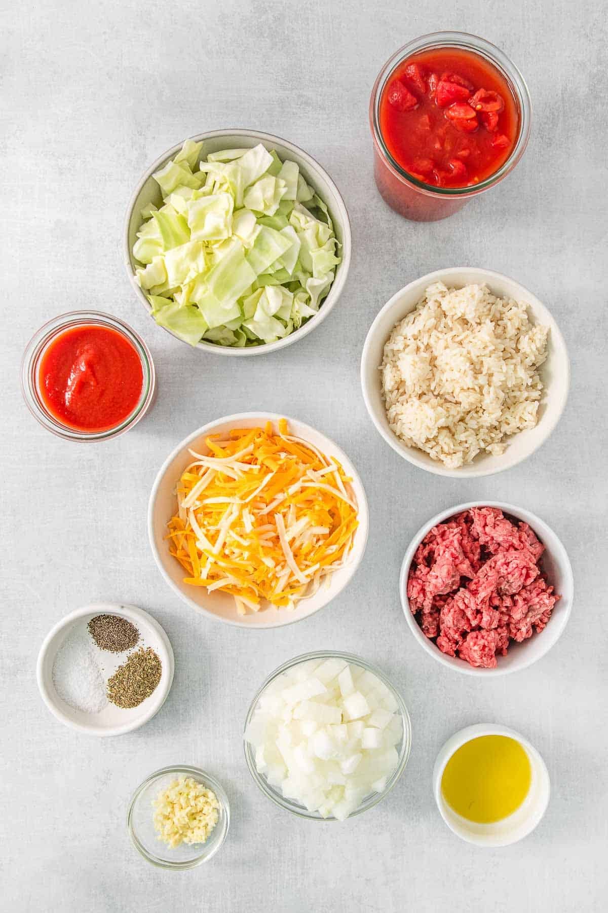 bowls containing ingredients for cabbage roll casserole - Olive oil, green cabbage, salt, ground beef, onion, garlic, tomatoes, tomato sauce, Italian dressing, black pepper, white rice and shredded cheese.