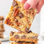Stack of three magic cookie bars on a white plate with one being picked up by woman's fingers.