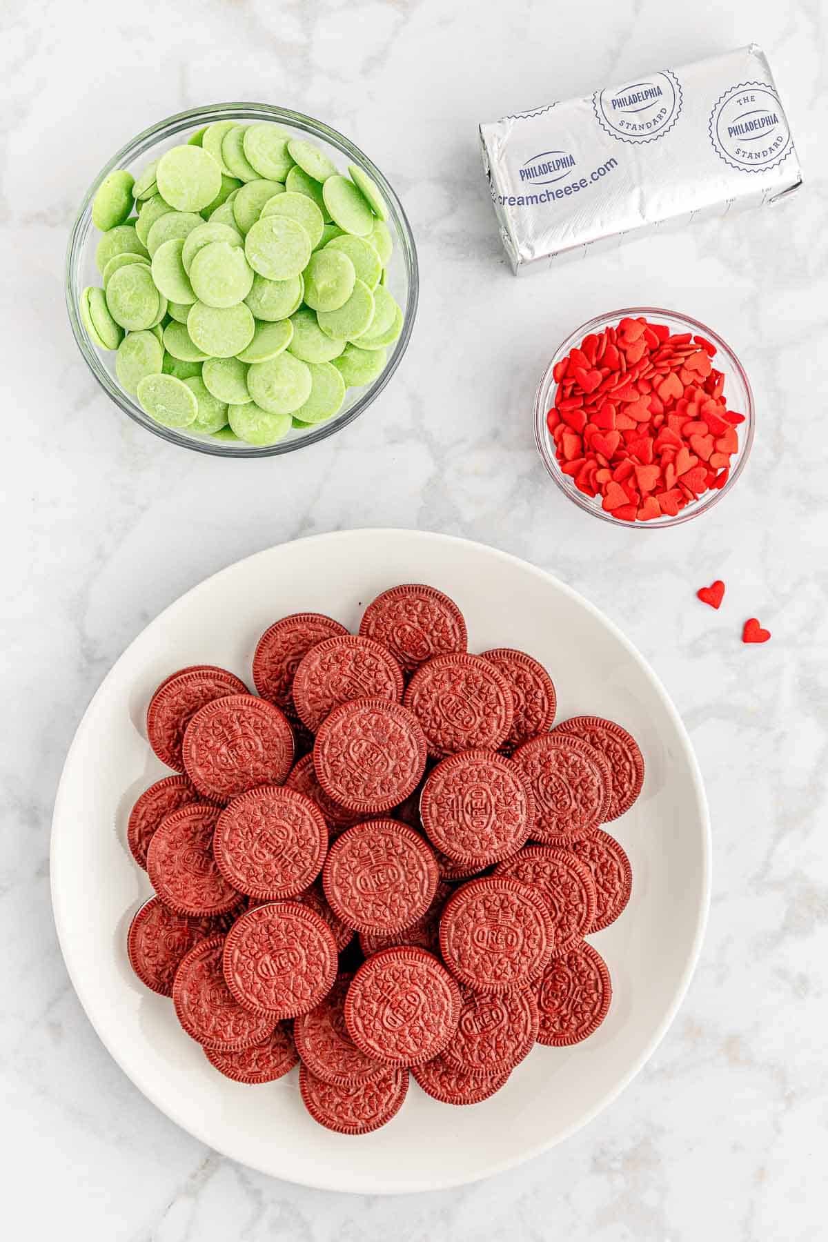 ingredients for grinch oreo cookie truffles on a marble countertop - white plate full of red velvet oreos, bowl of light green candy melts, red candy hearts and a block of cream cheese.