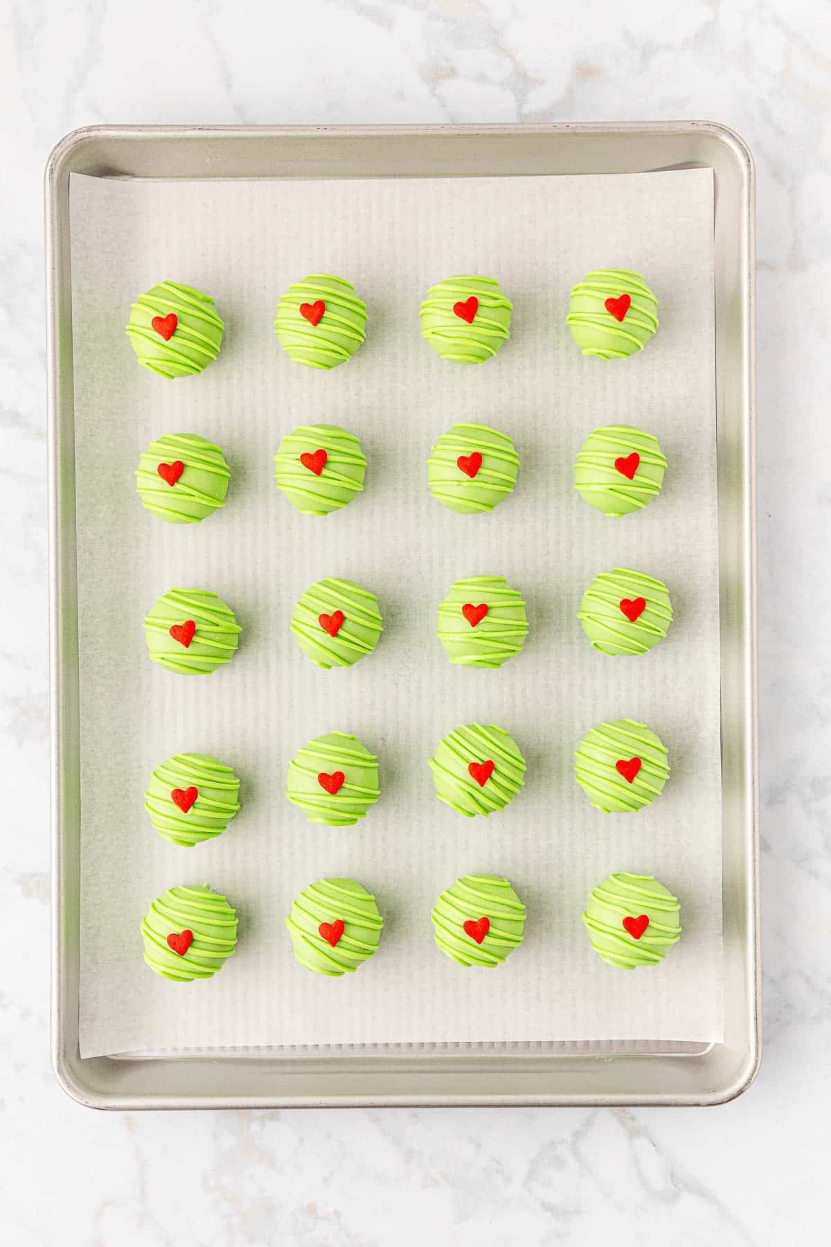 Green candy coated oreo balls with a red heart on each one on a baking sheet.