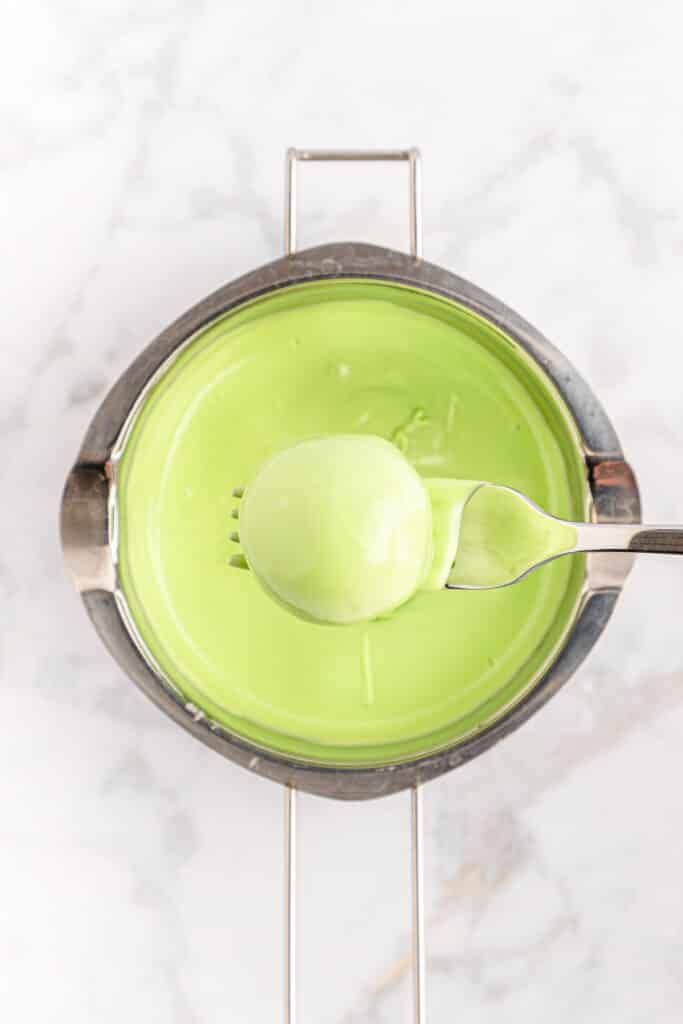 A fork holding a green candy coated ball over a in a pan of melted candy wafers.