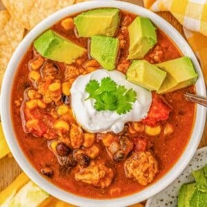 A white bowl of pumpkin chili topped with sour cream and avocados surrounded by tortilla chips.