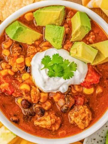 A white bowl of pumpkin chili topped with sour cream and avocados surrounded by tortilla chips.