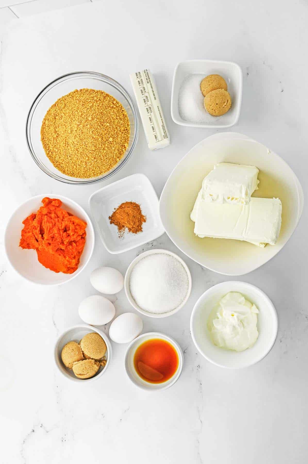 The ingredients for a pumpkin swirl cheesecake bars - Graham cracker crumbs, butter, sugar, brown sugar, pumpkin puree, pumpkin pie spice, cream cheese, sour cream, vanilla extract and eggs.