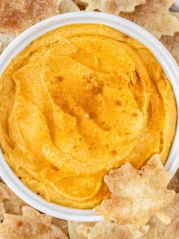 A bowl of pumpkin dip in a white bowl surrounded by cinnamon tortilla chips.